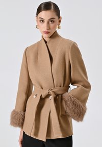 Cappotto avvolgente in misto lana color panna con vita cinturata, caratterizzato da morbidi dettagli in pelliccia sulle maniche e grandi bottoni per la chiusura.