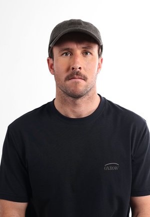 Homme avec une petite barbe et une moustache portant une casquette gris foncé et un t-shirt noir Oxbow, faisant face à un fond blanc uni.