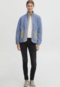 Fleecejacka i ljusblått, med beige detaljer och två framfickor. Ståkrage, långa ärmar och elastiska muddar. Matchad med svarta jeans.