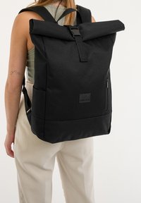 Person wearing beige pants and green sleeveless top with large black roll-top backpack secured by front buckle and side zipper pocket.