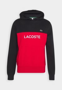 Hoodie med svart kropp och röd bröstpanel, stor vit "LACOSTE" text och logotyp på vänster sida. Tillverkad av bomullsblandning.