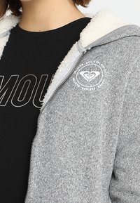 Felpa grigia con zip e colletto foderato in morbido pile; presenta un logo circolare sulla parte sinistra del petto e un tessuto texturizzato.