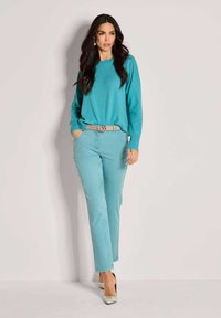 Turquoise trui met lange mouwen, gecombineerd met lichtblauwe broek en een beige riem. De schoenen hebben een patroon, wat de outfit benadrukt.