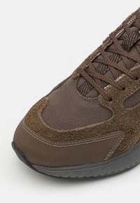 Brown sneaker featuring a blend of textured suede and mesh materials, with a curved toe and black rubber sole, detailed stitching visible.