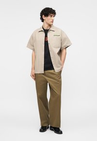Homme portant une chemise beige à manches courtes boutonnée par-dessus un tee-shirt graphique noir, pantalon marron à jambes larges et chaussures noires, debout devant un fond blanc.