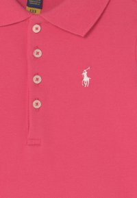 Różowa koszulka polo z fakturowanej tkaniny, z trzema guzikami i białym wyszywanym logo przedstawiającym konia z jeźdźcem na lewej piersi.