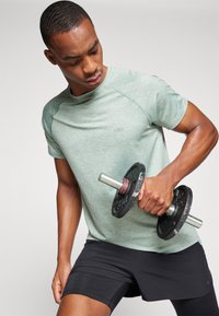Camiseta atlética de manga corta verde claro con un diseño texturizado, combinada con pantalones cortos negros. La persona sostiene un dumbbell negro.
