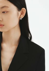 Pendientes de plata con una gema transparente facetada en el centro, rodeada de un diseño circular de metal texturizado. Lucidos por un modelo.