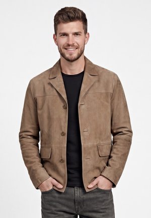 Jonge man die lacht, met kort bruin haar, draagt een beige suède jas over een zwart overhemd en donkergrijze spijkerbroek, staand met handen in de zakken.
