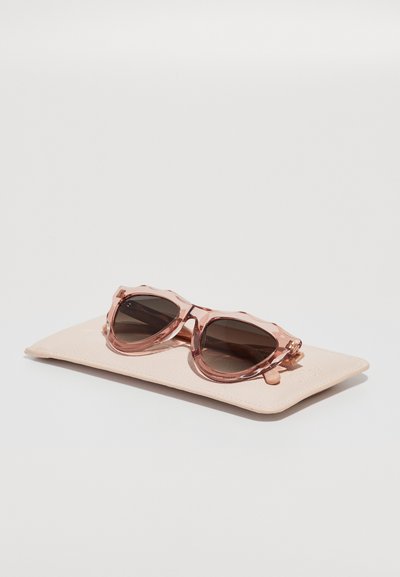 Lunettes de soleil teintées de rose avec une monture texturée et ondulée, reposant sur une étui doux rose clair. Les verres sont foncés avec un léger dégradé.