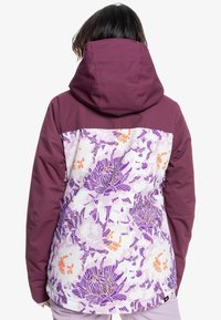 Kvinna som bär en lila jacka med huva, med enfärgade ärmar och övre delen av ryggen, samt en blommig mönstring i lila, orange och vitt på nedre delen av ryggen.