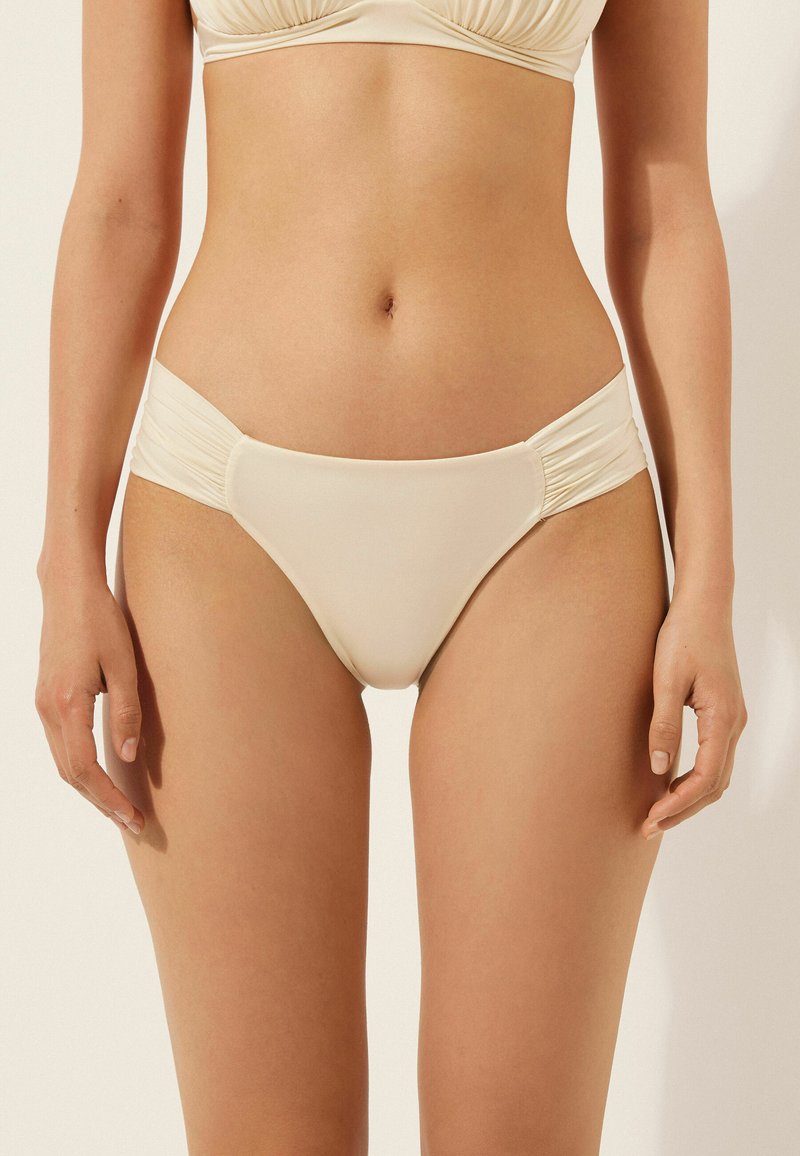 Helle beige Bikini-Brief mit Raffung an den Seiten, glattem Stoff, niedrigem Schnitt und minimaler Bedeckung.