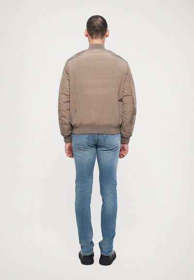 Jachetă bomber maro cu finisaj texturat, mâneci și tiv ribbed, asortată cu blugi slim-fit de culoare albastru deschis și pantofi negri, văzută din spate.