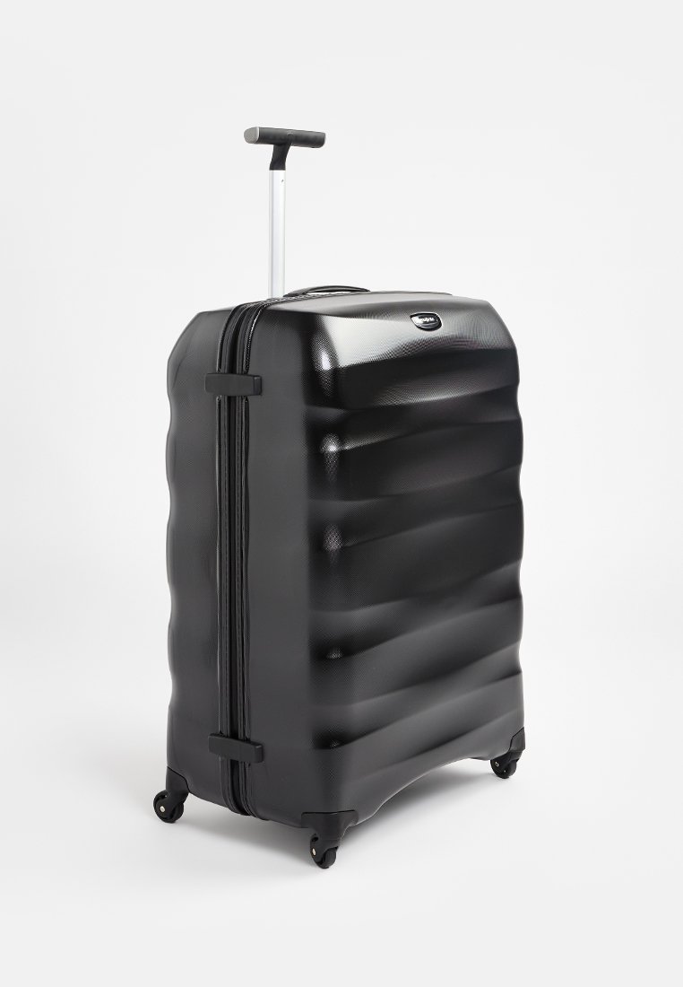 Schwarzer Hartschalenkoffer mit strukturiertem, wellenförmigem Design, ausziehbarem Griff und vier Rädern für Mobilität. Reißverschluss vorne.