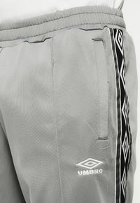 Šedé športové nohavice Umbro s čierno-bielymi geometrickými bočnými pruhmi a bočnou vreckou, nosené s bielym topom.