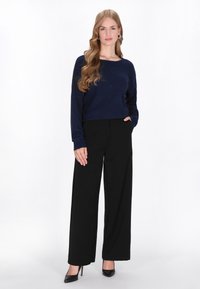 Granatowy sweter z dzianiny, czarne spodnie szerokie, czarne szpilki, modelka stoi z rękami w kieszeniach, gładka faktura, prosty design.