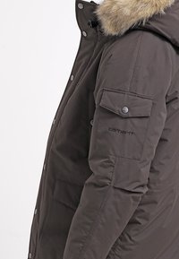 Parka marrone scuro con cappuccio foderato in pelliccia sintetica, chiusura a bottoni, tasche laterali e logo ricamato sulla manica. Tessuto con una texture liscia.