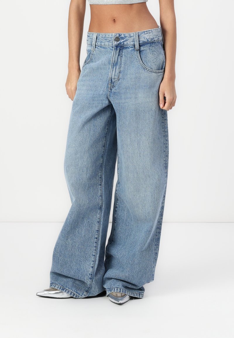 Jeans largi de culoare albastru deschis, cu talie înaltă, închidere cu nasture, textură de denim uzată și buzunare laterale adânci. Accentuate cu pantofi cu toc argintii.