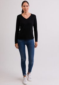 Schwarzer, langärmeliger V-Ausschnitt-Pullover kombiniert mit enganliegenden, mittelblauen Jeans und weißen Turnschuhen. Glatte Textur, schlichtes Design und enganliegende Form.