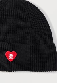 Gorro de punto negro con textura acanalada, con un parche de corazón rojo que dice "HUGO" en texto blanco.