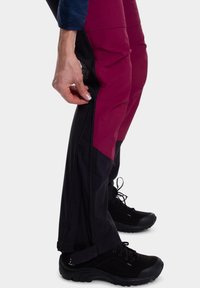 Maroon and black zippered pants with a slim fit and textured fabric, featuring side zippers and paired with black hiking shoes.