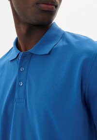 Polo de algodón azul con cuello clásico, tapeta de tres botones y detalle sutil del logo en el pecho. Textura suave, ajuste entallado.