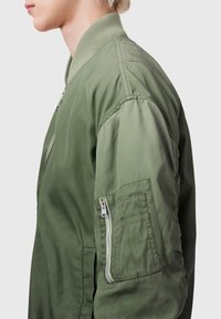 Olive green bomber jacket with a ribbed collar, zippered chest pocket, and textured fabric. Features a smooth finish and relaxed fit.