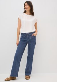 Bílý strukturovaný krátký rukávový top v kombinaci s volnými modrými denimovými kalhotami se stahovacími šňůrkami v pase a postranními kapsami, obutý s hnědými sandály.