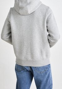 Felpa grigia in pile con cappuccio curvo, polsini e orlo a costine. Vestibilità ampia, tessuto testurizzato, abbinata a jeans blu chiaro con tasche posteriori.