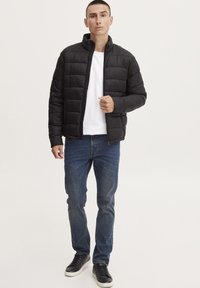 Jachetă neagră tip puffer cu design matlasat, guler înalt și închidere cu fermoar în față. Potrivită cu blugi albaștri și adidași negri. Stil simplu și funcțional.
