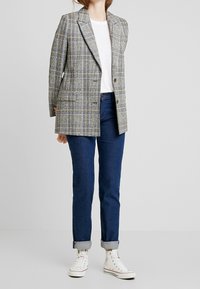 Blazer de cuadros gris con solapas de pico, combinado con una camiseta blanca y jeans de un azul oscuro, complementado con puños enrollados y zapatillas blancas.