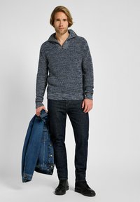 Suéter de punto azul marino con un cuello alto, patrón texturizado y puños de canalé, combinado con jeans oscuros y sosteniendo una chaqueta de mezclilla.