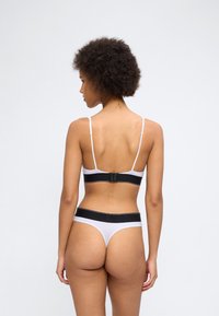 Γυναίκα με σγουρά μαλλιά, εμφανιζόμενη από πίσω, φοράει λευκά και μαύρα εσώρουχ α μπικίνι μπροστά σε απλό φωτεινό φόντο.