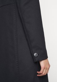 Manche de manteau noire avec des coutures visibles et un seul bouton noir près du poignet, avec une main suspendue détendue à côté.