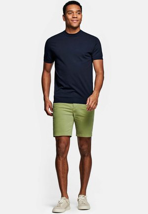 Man loopt naar voren en draagt een marineblauw shirt met korte mouwen, lichtgroene korte broek en beige sneakers tegen een effen witte achtergrond.