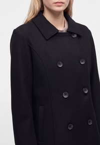 Jeune femme portant un manteau noir croisé avec de gros boutons et un large col, montrée du torse au cou.