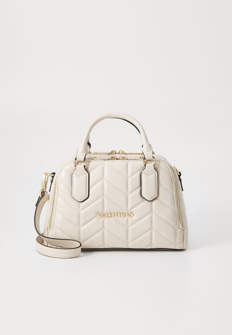 Beige laukku, jossa on tikattu pinta, kultaiset yksityiskohdat, ylhäältä kannettavat kahvat ja irrotettava olkahihna. sisältää kohokuvioidut brändäykset ja vetoketjumainoksia.