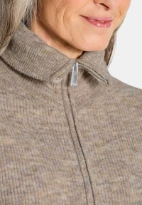 Beige zip-up Sweater mit hohem Kragen, gerippter Textur und silbernem Reißverschluss. Der Stoff zeigt subtile Farbvariationen.