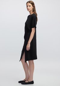 Robe noire à manches courtes, longueur genou, avec une fente sur le côté et un détail noué à la taille. Assortie à des chaussures noires plates.
