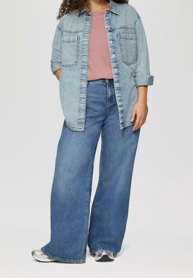 Personne portant une veste en jean clair sur un t-shirt rose et un jean bleu à jambes larges avec des baskets argentées, debout devant un fond uni.