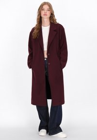 Donna con capelli mossi, che indossa un lungo cappotto borgogna su una maglietta corta bianca, jeans a gamba larga scuri e sneakers bianche, in piedi con le mani nelle tasche.