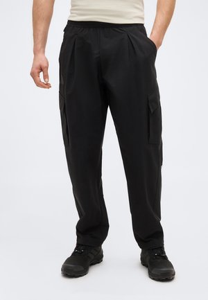 Hombre vistiendo pantalones cargo negros con múltiples bolsillos, combinados con zapatos de senderismo negros, de pie frente a un fondo blanco liso.