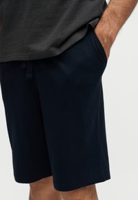 Homme portant un short bleu marine foncé avec poches latérales, main dans la poche droite, associé à un t-shirt gris foncé.