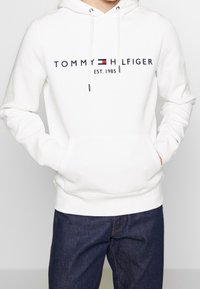 Biały hoodie z regulowanymi troczkami, przednia kieszeń oraz logo "TOMMY HILFIGER EST. 1985" w granatowym i czerwonym kolorze. Materiał mieszanka bawełny.