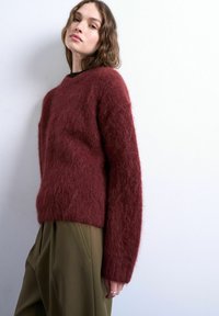 Femme portant un pull bordeaux en laine pelucheuse et un pantalon taille haute vert olive, debout contre un mur blanc, regardant légèrement vers le haut.