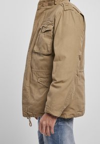 Mężczyzna w beżowej kurtce typu cargo z wieloma klapkowymi kieszeniami, w jasnoniebieskich dżinsach, stojący na tle jednolitego szarego tła.