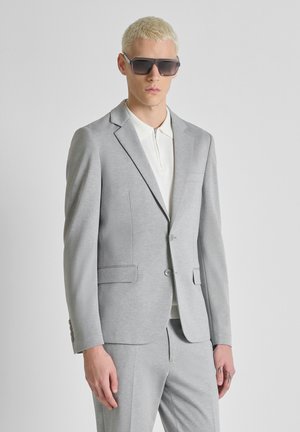 Uomo con capelli biondi corti che indossa grandi occhiali da sole neri e un completo grigio chiaro con una camicia bianca con cerniera su uno sfondo neutro.