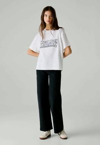 Valge puuvillane T-särk, millel on sinise värviga tekst "Red Bull Racing", koos mustade laiemate pükstega ja valgete tossudega. Lihtne disain, vabalt istuv.