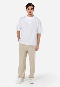 T-shirt en coton blanc à col rond et manches courtes, avec du texte noir au centre. Associé à un pantalon beige et des baskets blanches.