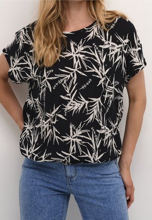 Kvinde iført en sort kortærmet top med hvidt botanisk print kombineret med blå denimjeans, stående foran en ensfarvet baggrund.